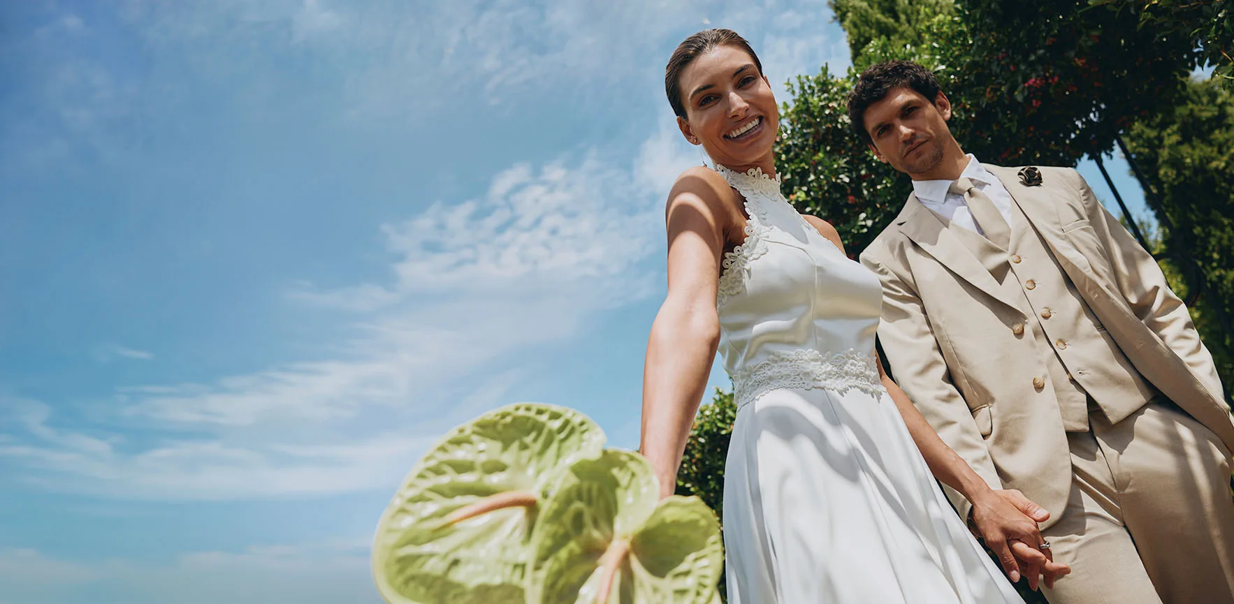 Jeunes mariés par une belle journée ensoleillée : la mariée porte une robe blanche, le marié un costume de couleur beige.